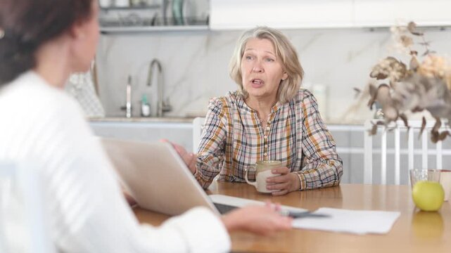 Positive old woman sitting at the kitchen-table face to face with middle-aged female agent demonstrating something on her laptop