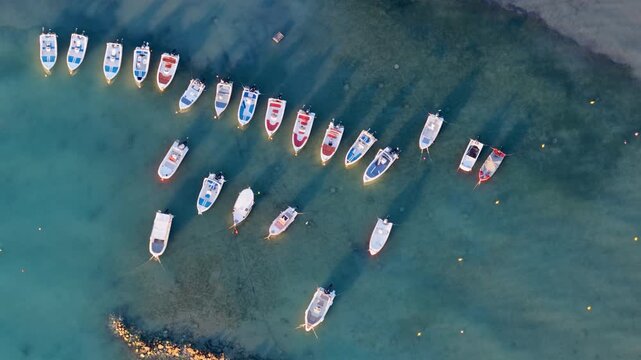 Drone close up top down view tracking along row of small boats anchored in crystal clear turquoise water, Sidari beach, Corfu, Greece. Aerial summer holiday marine travel background.
