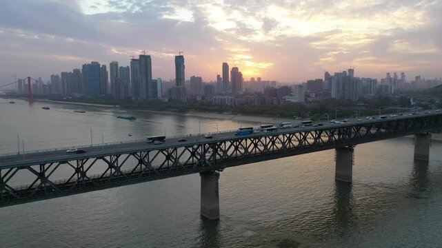 Wide sunset aerial shot of traffic crossing the steel truss First Wuhan Yangtze River Bridge over the Yangtze River in Wuhan, Hubei, China, framed by an expansive modern city skyline at dusk.