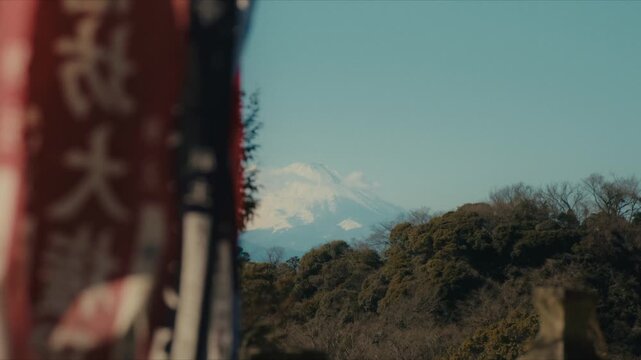 A distant view of snow-capped Mount Fuji seen from the Hansobo Shrine observation deck atop Kencho-ji temple in Kamakura, with traditional red and white shrine banners in the foreground.