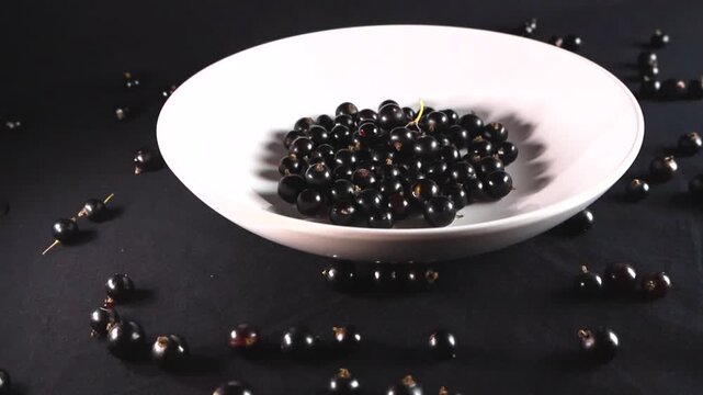 Close up view of fresh and juicy blackcurrant berries in a white plate, with other berries scattered on a dark background, showing the concept of healthy eating and organic food