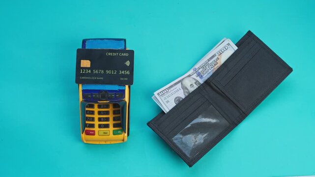 Credit card sits on payment terminal next to an open wallet with cash on turquoise background