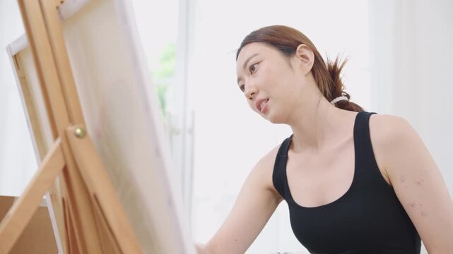 Beautiful, happy Asian Japanese woman artist in black tank top painting watercolor with paintbrush on canvas on wooden easel in clean white bright home studio room with calm, relaxation and happiness