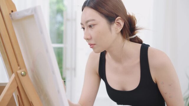 Beautiful, happy Asian Japanese woman artist in black tank top painting watercolor with paintbrush on canvas on wooden easel in clean white bright home studio room with calm, relaxation and happiness