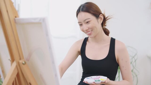 Beautiful, happy Asian Japanese woman artist in black tank top painting watercolor with paintbrush on canvas on wooden easel in clean white bright home studio room with calm, relaxation and happiness