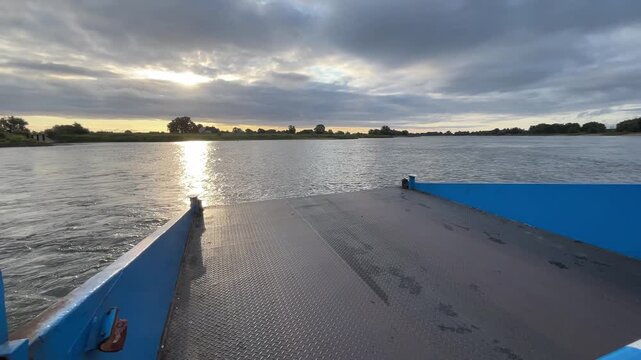 Open Vehicle Ferry Deck During Evening River Crossing with Sun Reflection on Calm Water Beneath Dramatic Cloudy Sky and Wide Riverside Landscape