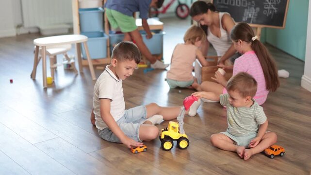 Attentive two boys sitting on floor and playing with cubes and cars in modern childcare center, other children in the background