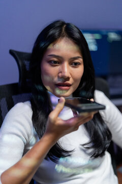 Asian woman tech worker speaking to voice assistant on smartphone at night, using AI voice search and speech interface with code light reflecting on her face