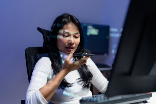 Asian woman developer speaking voice command to smartphone while working at computer workstation at night, using AI to help her workflow with code light on her face