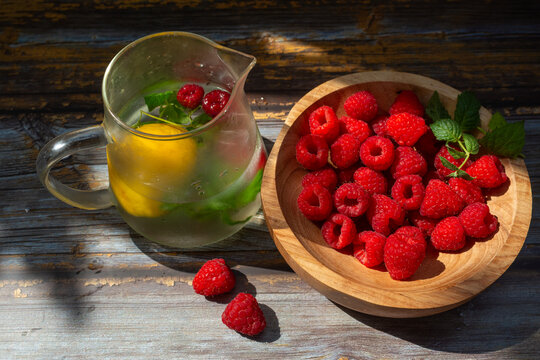 Fresh raspberries, Rubus idaeus, served beside a glass pitcher of cold infused water with lemon, peppermint leaves, and floating berries. Rustic summer food composition promoting hydration, healthy ea