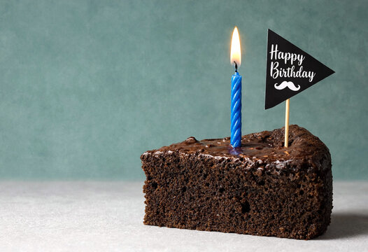 Delicious chocolate birthday cake slice with a single lit blue candle and happy birthday flag