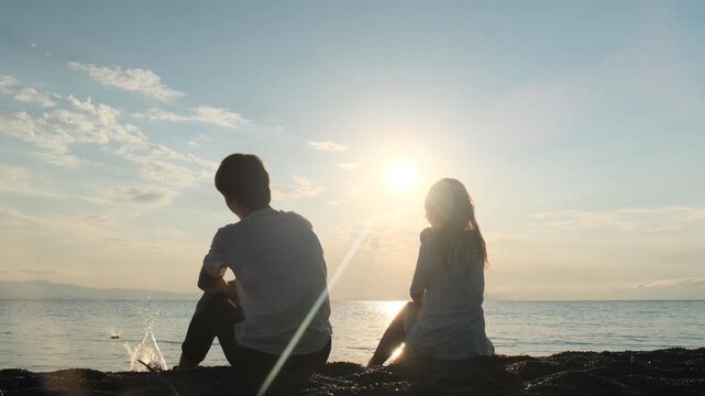 Rear view of a teenage boy and girl throwing stones into the sea together at sunset. First love, sibling friendship, quality time outdoors and carefree summer moments.