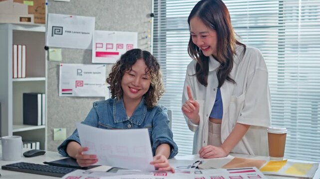 Two Asian Designer woman meeting brainstorming ideas about design logo,discuss on tablet and paper,female colleagues working together design banner logo in office.Graphic Design and Designer concept
