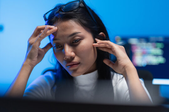 Woman software developer leans toward a screen with hands at her head, code visible on a monitor behind her in a blue lit room during a late night work shift.