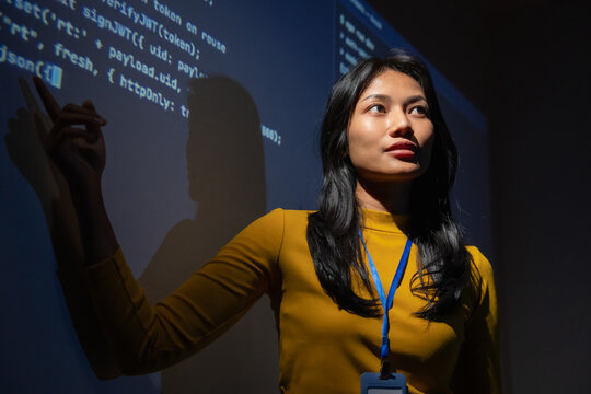 Woman software engineer with lanyard points at authentication code projected in a dark room, reviewing session token and login security logic during a technical walkthrough.