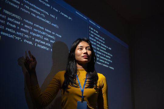 Woman software developer raises a finger as an idea comes to mind while reviewing projected code and server logs in a dark room, focused on solving a technical problem.