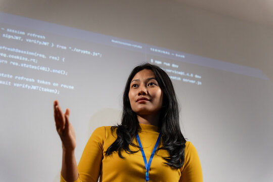 Professional woman developer presenting code on projection screen in tech training environment.  confident expression, open hand gesture indicating active teaching or presentation.