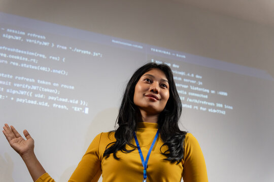 Smiling woman developer in yellow top with lanyard stands with open palm gesture, programming code projected behind her, conveying confidence in a technology career.
