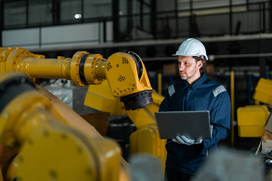Professional male engineer in a blue uniform and hard hat using a laptop to inspect and program a yellow industrial robotic arm in a modern smart factory, focusing on industry 4.0 automation.