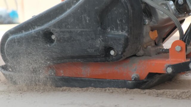 Close up of a plate compactor compacting a sand base before paving stone installation. Gasoline powered construction equipment prepares the foundation for sidewalk construction.