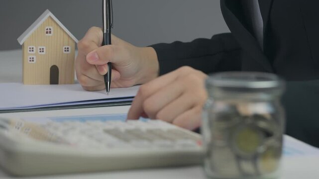 Person planning financial paper goal, writing notes near calculator device, saving coins beside wooden house model for future home ownership