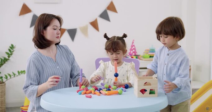 Asian female kindergarten teacher supervising toddler boy, toddler girl assembling colorful construction toy on kids table, bright playroom decor, preschool learning, social skill, imagination.