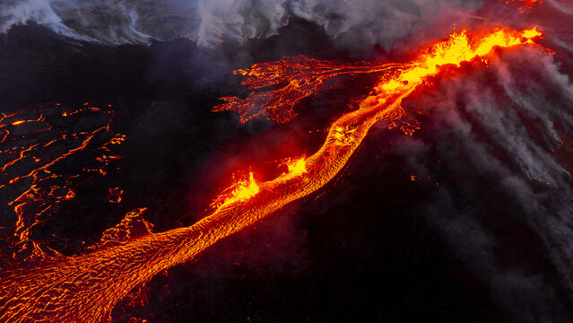 Aerial view of the Litli-Hrutur volcanic eruption with glowing lava flowing across the dark landscape and plumes of volcanic smoke rising into the air in Grindavik, Grindavikurbaer, Iceland.