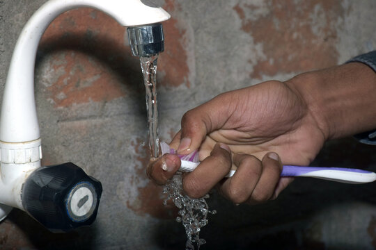 Close-up view of white and purple toothbrushes being washed under running water from a plastic faucet in front of a brick wall