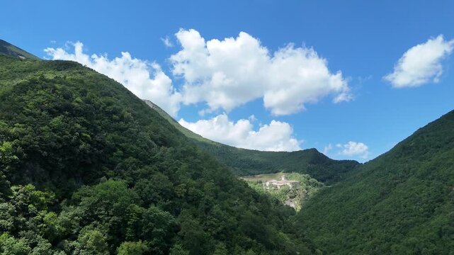 Paesaggio montano con due rilievi verdi ricoperti da foresta, valle chiara centrale e cielo azzurro con nuvole bianche