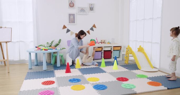 Asian female preschool teacher reaches to adjust cone lane, preschool boy walks near slide, preschool girl runs across balance discs, daycare playroom agility drill, coordination practice, bright room
