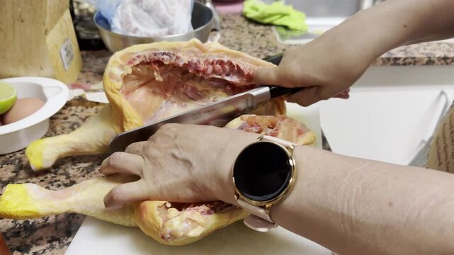 A woman's hands using a large butcher's knife to cut up a chicken on a cutting board