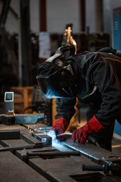 Vertical image of young adult welder in protective mask and gloves welding metal parts in workshop, representing industrial manufacturing and vocational training school promotions
