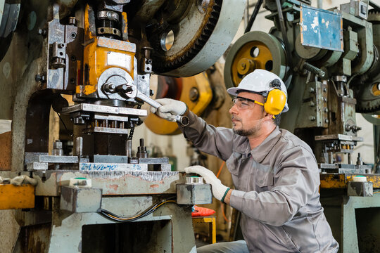 Male engineer kneeling at a power press reaching up with a wrench toward the die head in a metal stamping factory