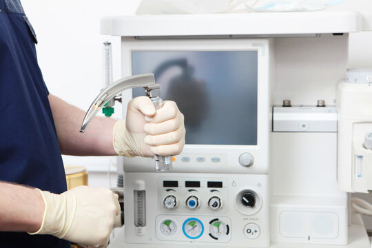 Doctor's hand in sterile gloves holds laryngoscope against background of modern anesthetic device in operating room
