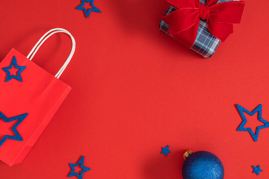 Christmas Shopping and Gift Flat Lay Background on Red Backdrop with Copy Space. Red Paper Bag, Plaid Present Box with Velvet Bow, Blue Bauble and Glitter Stars Top View. Festive Winter Holiday Concep