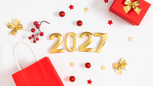 Happy New Year 2027 Festive Flat Lay Banner with Golden Numbers. Red Shopping Bag, Christmas Gift Box, Bows and Confetti Stars on White Background. Winter Holiday Shopping and Celebration Concept.
