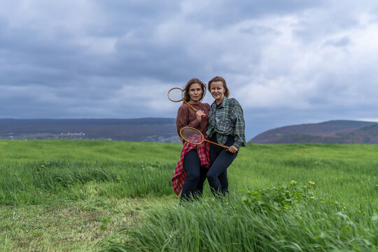 Active young women posing with wooden badminton rackets in a lush green meadow landscape during an outdoor leisure activity