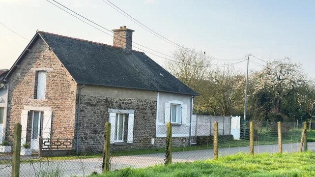 Stone village house with fenced yard beside quiet rural road, spring trees in bloom