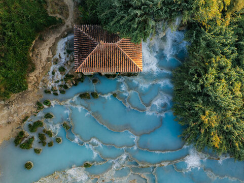 Aerial view of the Cascate del Mulino thermal springs featuring turquoise tiered pools, a historic stone mill building, and lush surrounding vegetation in Saturnia, Tuscany, Italy.