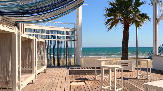 Beautiful empty beach lounge area with wooden deck and palm tree facing ocean waves on sunny day.