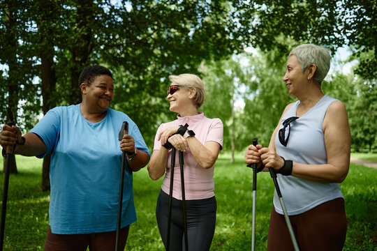 Diverse senior women holding Nordic walking poles in park, smiling and talking. Image represents active aging and wellness, ideal for senior healthcare and fitness marketing campaigns