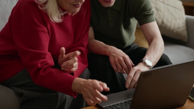 Caucasian senior woman and Caucasian young adult man sitting on sofa, looking at laptop screen and discussing content. Clip is suitable for promoting digital literacy programs for seniors