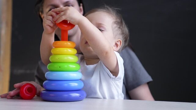 Toddler stacks colorful toy ring on table. Calm mother watches child play. Boy puts bright plastic ring on stacking toy. Focused toddler learns early development skill. Mother helps tiny toddler.