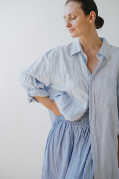 Woman In Light Blue Oversized Linen Shirt And Smocked Waist Skirt Posing Standing Against White Wall