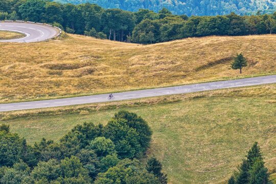 Aerial view of a cyclist riding along a winding mountain road on Grand-Ballon surrounded by grassy slopes and patches of forest in Soultz-Haut-Rhin, Grand Est, France.