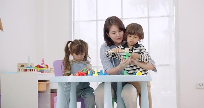 Asian female preschool teacher holding toddler boy, helping place peg stack piece, toddler girl playing alone at table, daycare behavior support, emotional regulation, learning through play.
