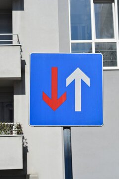 A square blue traffic sign indicating priority over oncoming traffic with a white arrow pointing up and a red arrow pointing down stands in front of a residential building facade