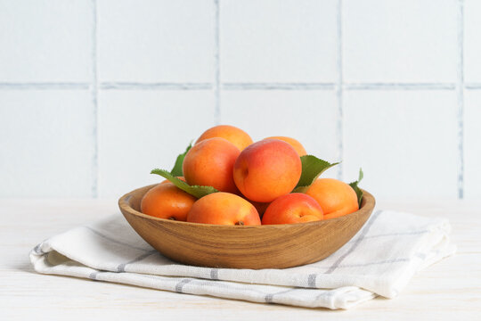 Fresh ripe apricots arranged in a wooden bowl on white kitchen table and tiled wall background with copy space. Summer stone fruits with green leaves, rustic home kitchen setting, healthy food