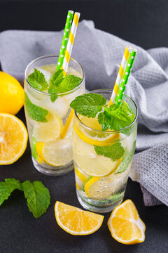 Close up of refreshing cold icy beverage, homemade lemonade with fresh lemon slices, mint leaves, ice cubes wtih soda water and condensation in glasses on black textured background