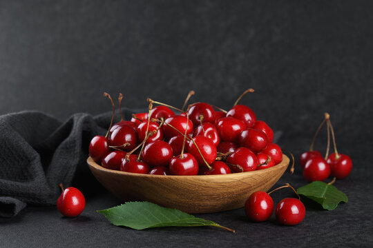 Fresh ripe red sweet cherries in wooden bowl on dark textured stone background with green leaves and black linen towel. Summer fruit, berry, harvest, healthy organic food